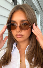 Close-up of brown oval rimless sunglasses with gold metal bridge and temple details, OTRA logo on lens