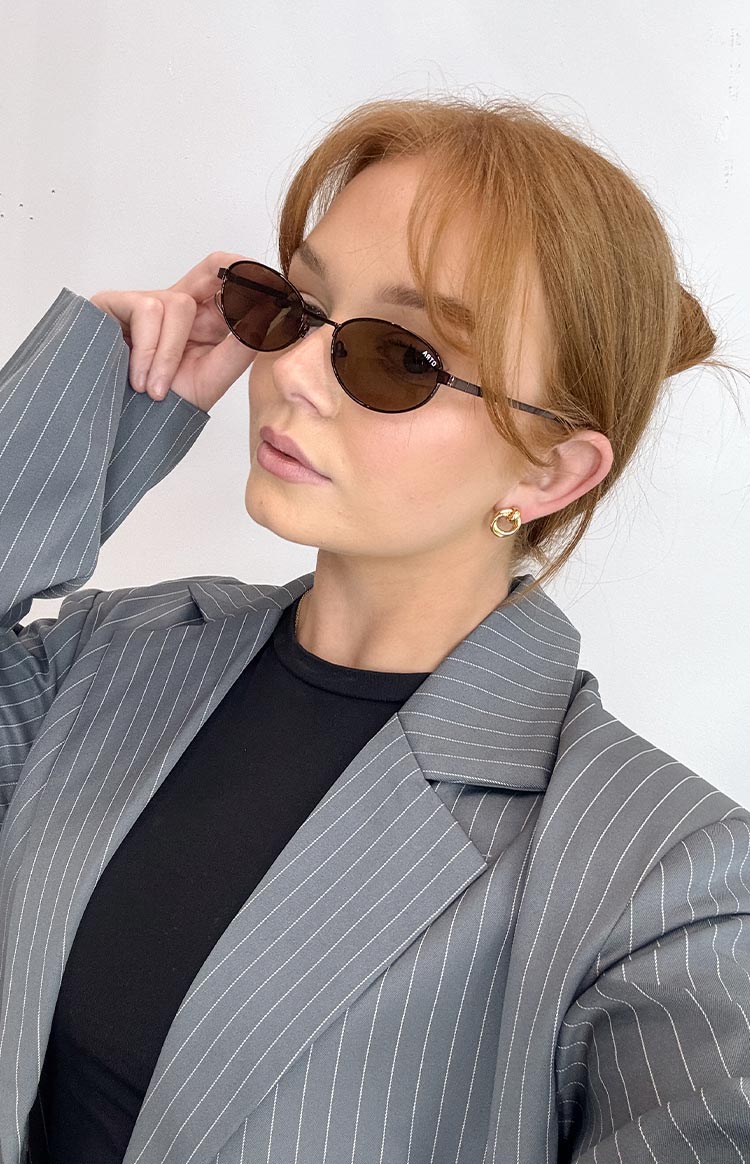 Brown oval-framed sunglasses worn with a grey pinstripe blazer and a black top.