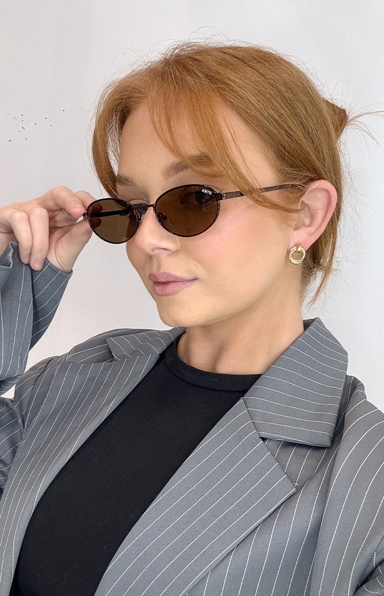 Brown oval-framed sunglasses, styled with a grey pinstripe blazer and a black top.