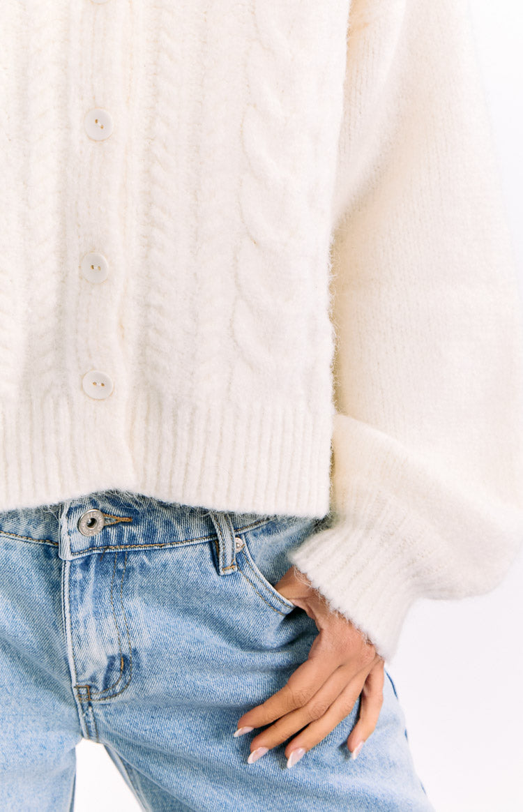 Close-up of the cream cable knit cardigan showing the button placket, ribbed hem, and sleeve cuff