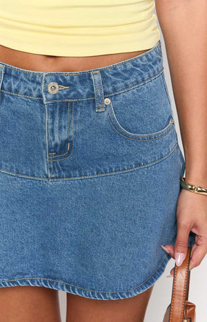 Close-up of the mid-wash denim mini skirt showing the button closure, zipper fly, front pockets, and horizontal seam detail