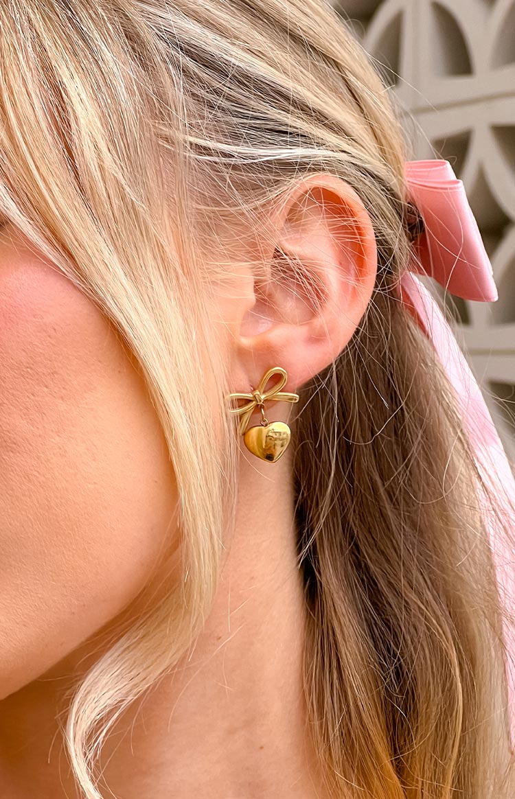 Close-up of a gold bow stud earring with a dangling polished heart charm