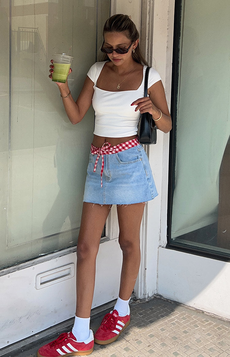 Light blue denim low rise mini skirt with an A-line cut, raw hem, and red gingham waist tie