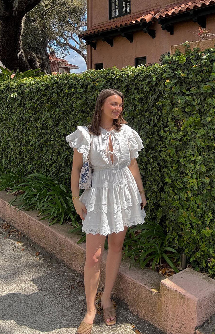 White mini dress with a V-neckline and tie detail, short flutter sleeves, pintuck bodice, and tiered skirt with eyelet trim