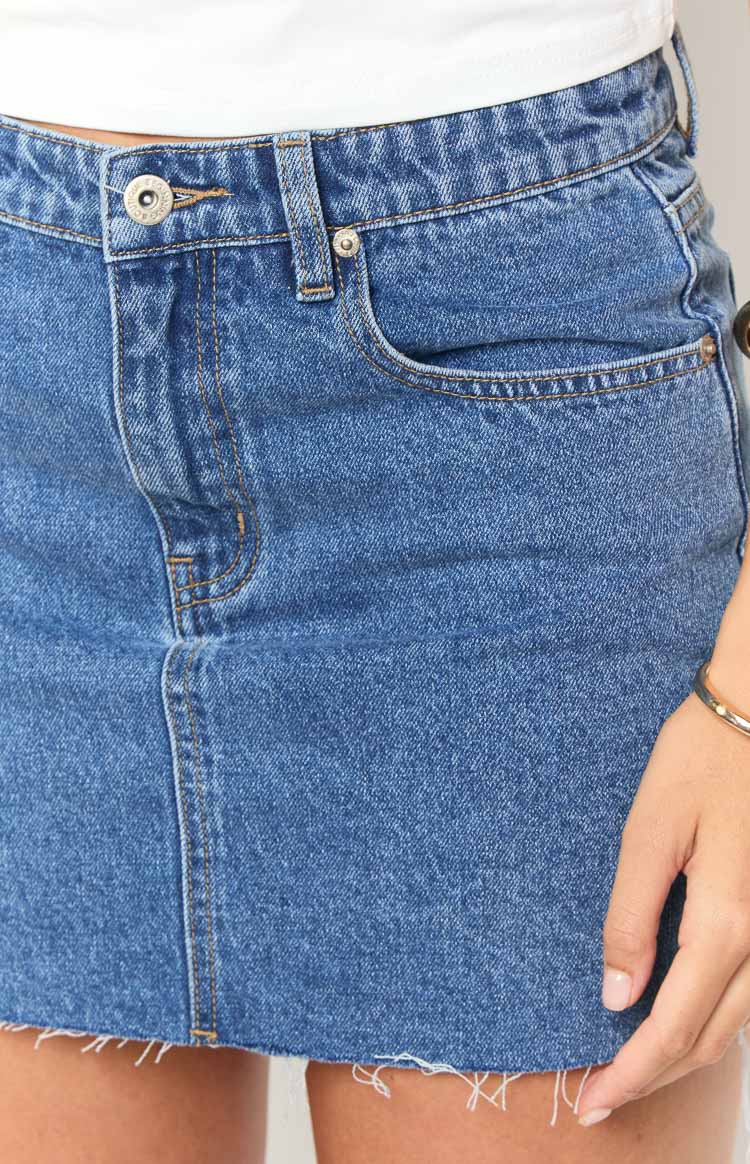 Close-up of the mid-wash denim mini skirt's front, highlighting the button closure, belt loops, and frayed raw hem