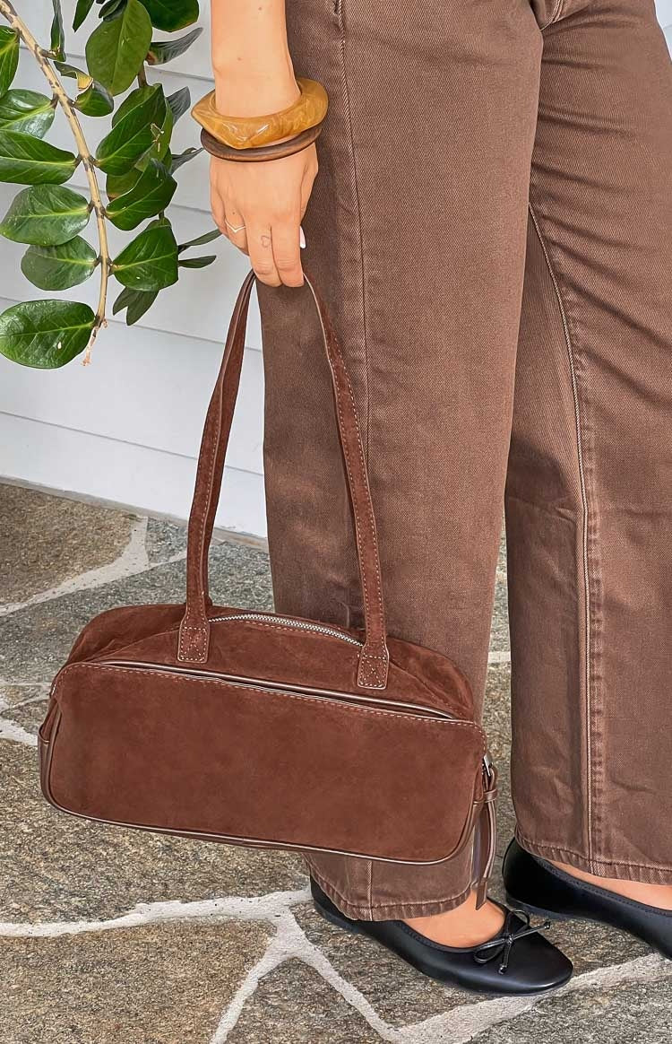 Close-up of a brown suede baguette bag showing the silver top zipper and stitched shoulder straps