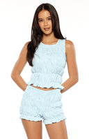 A lifestyle shot of our model wearing a light blue form-fitting top. This sleeveless top features all-over vertical smocking and horizontal gathered tiers ending in a ruffled hemline.