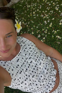 A close-up of a sleeveless, white and black polka dot crop top featuring a round neckline and a heavily smocked bodice with a ruffled hem.