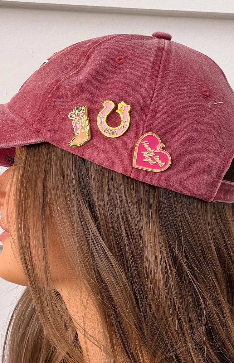 Side view of the three western pins on a maroon cap: a cowboy boot, a lucky horseshoe, and a heart