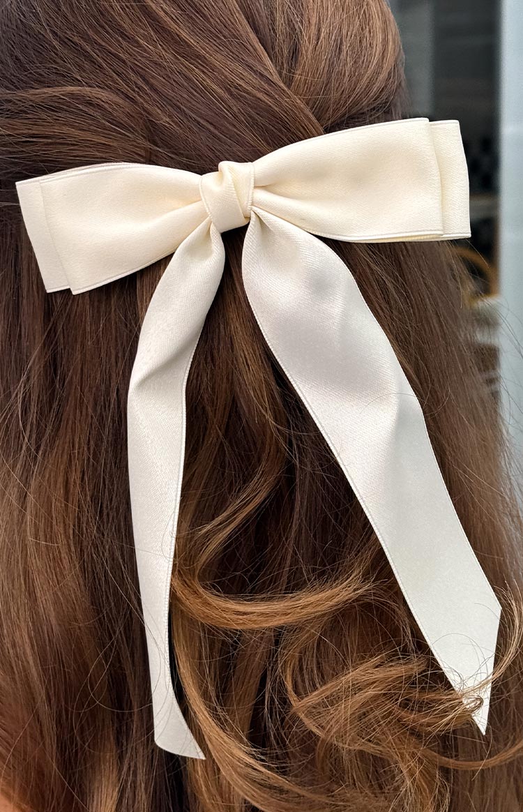 A close-up of a cream satin bow hair clip with long tails, securing a half-up hairstyle.