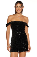 Our model wearing a form-fitting off-the-shoulder black mini dress. The entire dress is covered in shimmering black sequins. The neckline features small ruched off-the-shoulder straps.