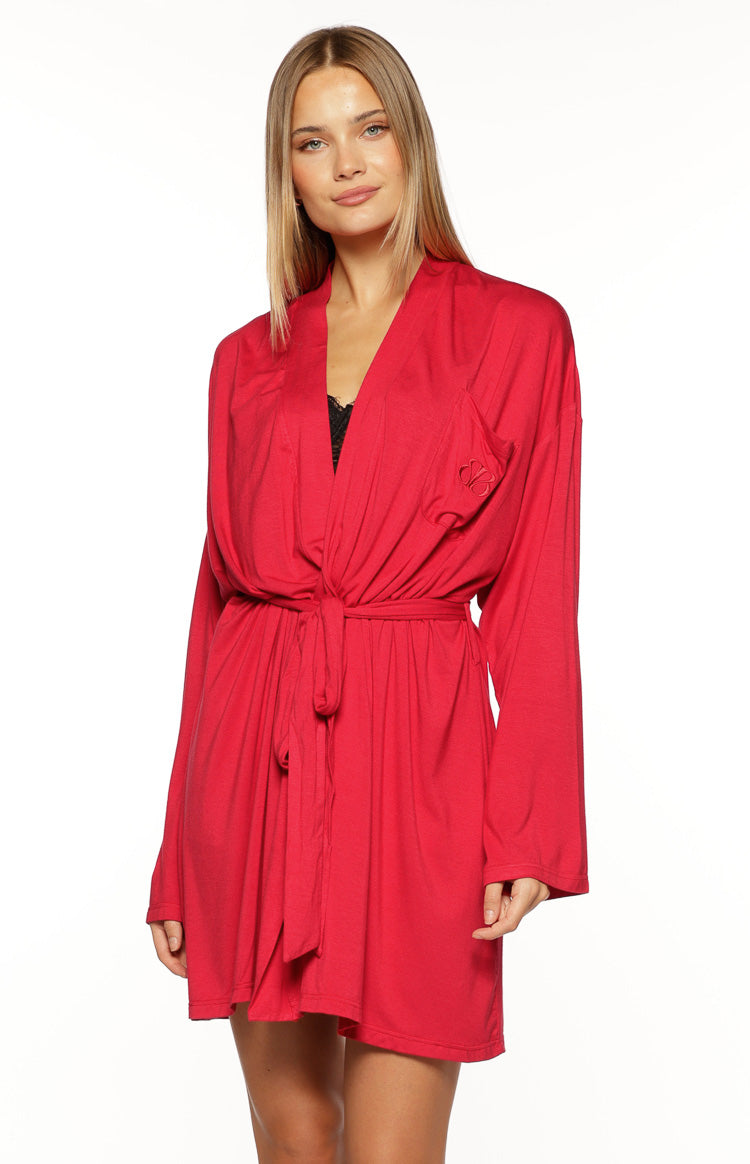 A close-up of or mode shows her wearing a short long-sleeve robe a scarlet red jersey knit fabric. This garment features a wrap front with a V-neckline a gathered waist and is tied with a thick matching fabric sash. Subtle stitch detailing is visible on the chest.