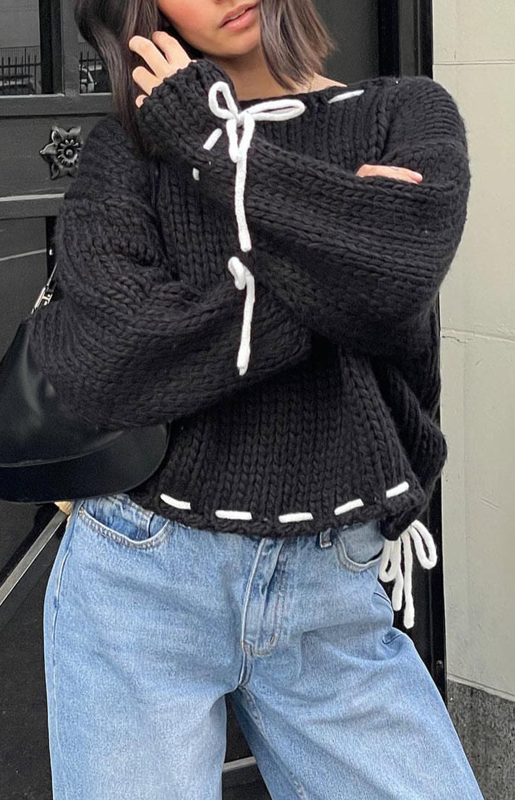 Close-up of a black chunky knit sweater with white lacing details and bows on the sleeves and hem
