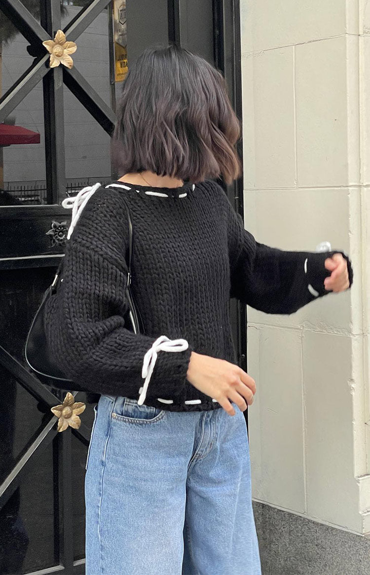 Side view of a black chunky knit sweater with white tie details and white stitching on the neckline and hem