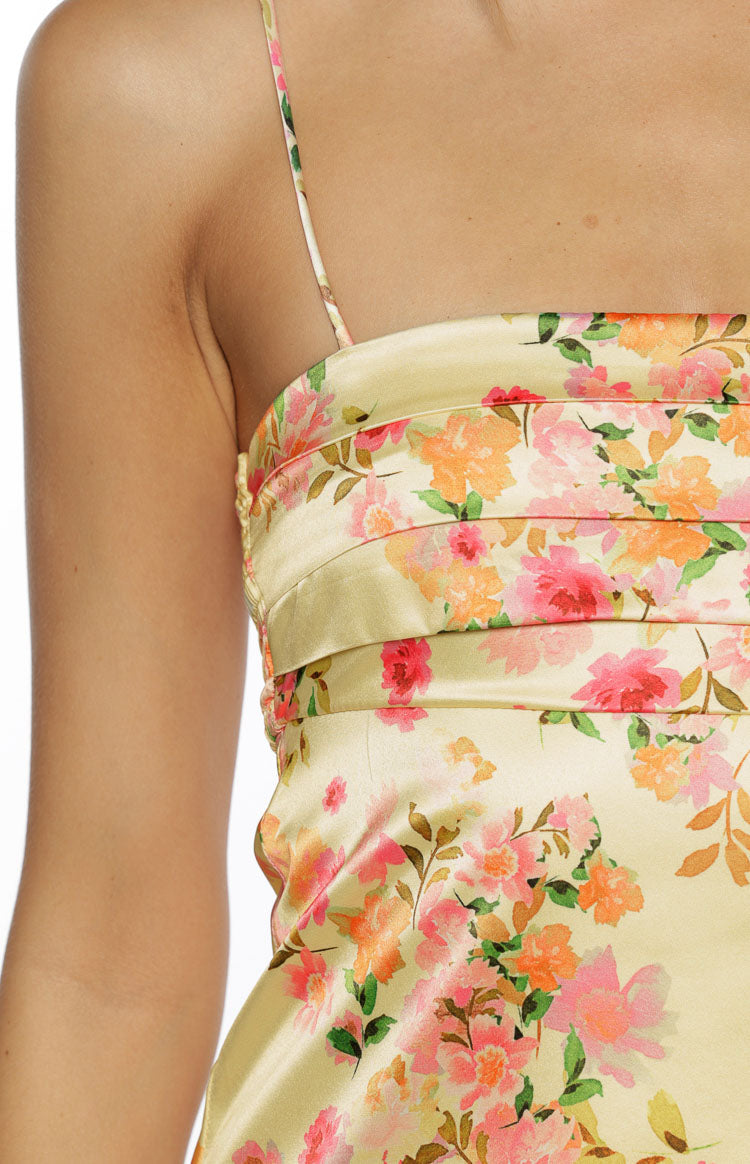 A tightly cropped close-up focuses on the bust of a light yellow satin dress. This mini dress fabric patterned with a dense print of pink coral and orange flowers features several horizontal pleats across the bustline creating a textured band detail. A single thin spaghetti strap is visible near the shoulder.