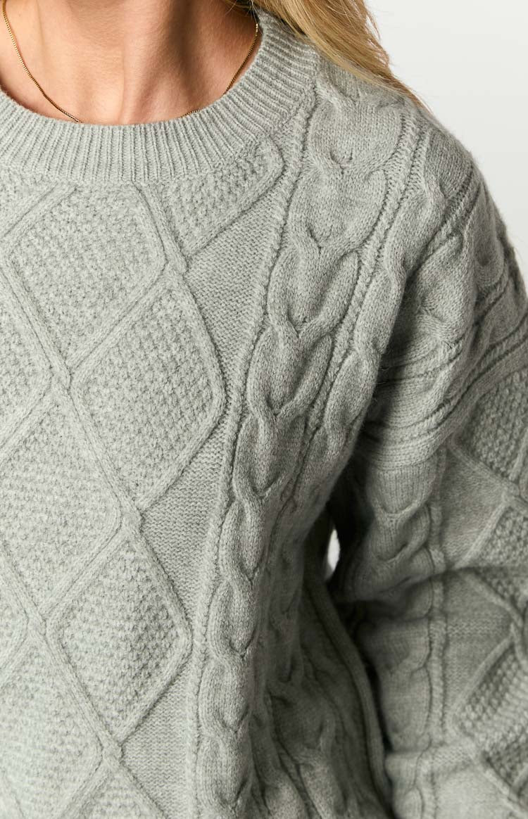 Close-up of the grey cable knit sweater showing the ribbed crew neck and textured diamond and braided patterns