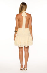 Rear view of a cream halter neck mini dress with an open back, tie detail, ruched waist, and tiered ruffle skirt