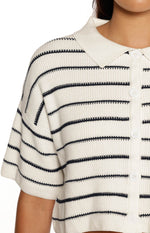 A close-up image of a cropped cream-colored knit polo shirt featuring thin horizontal navy blue stripes. The top has short sleeves with dropped shoulder seams a white fabric collar (contrasting slightly with the knit body) and a central button placket with large round white buttons. The knit texture is clearly visible.