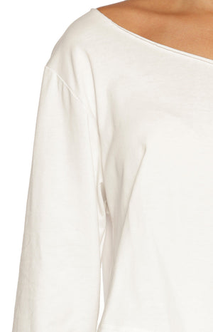 A cropped image focuses on the upper right shoulder and arm of our model wearing a simple white long-sleeve top. The garment features an extra-wide raw-edge neckline that sits off the shoulder exposing the collarbone and upper chest. The fabric appears to be a soft lightweight cotton.