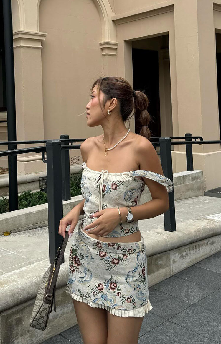 Side view of a beige tapestry mini skirt with floral pattern, low-rise fit, hip cutout, and ruffled hem