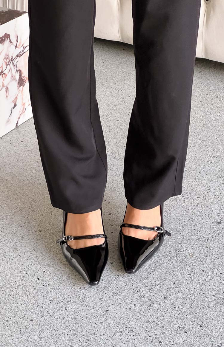 Close-up top view of black patent heels featuring a pointed toe and a delicate strap with a buckle