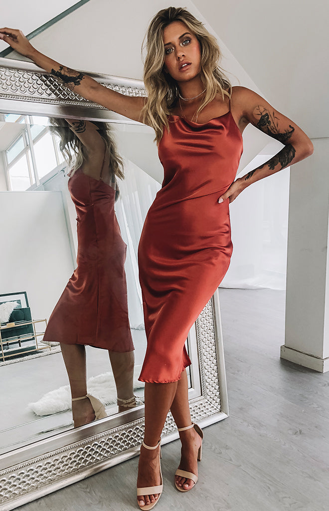 Rust satin midi dress with a flowing silhouette, cowl neckline, and delicate spaghetti straps